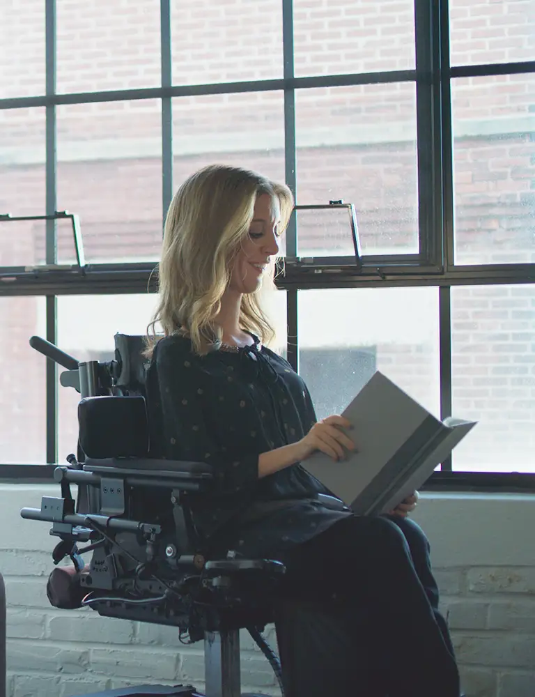Frau mit SMA sitzt in ihrem Rollstuhl am Fenster. 