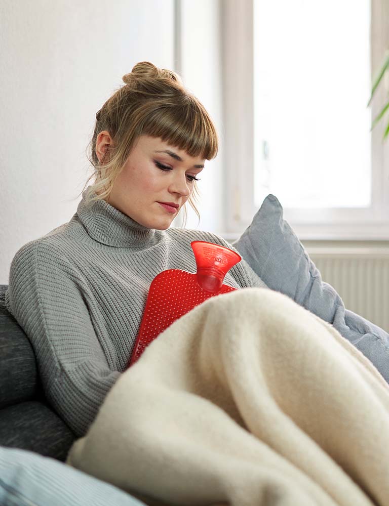 Gina sitzt mit ihrer Wärmflasche auf der Couch, denn sie leidet an einer CED (chronisch entzündlichen Darmerkrankung). 